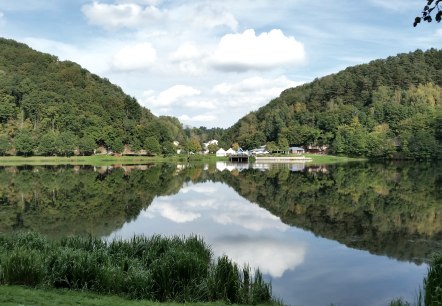 Panorama du lac de barrage de Bitburg avec des collines boisées, des bâtiments sur la rive et un reflet clair dans l'eau sous un ciel bleu., © TI Bitburger Land Panorama du lac de barrage de Bitburg avec des collines boisées, des bâtiments sur la rive et un reflet clair dans l'eau sous un ciel bleu., © TI Bitburger Land