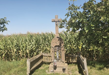 A stone cross with a figure of Jesus stands in front of a cornfield, surrounded by trees and a wooden fence. The sky is clear and blue., © TI Bitburger Land A stone cross with a figure of Jesus stands in front of a cornfield, surrounded by trees and a wooden fence. The sky is clear and blue., © TI Bitburger Land