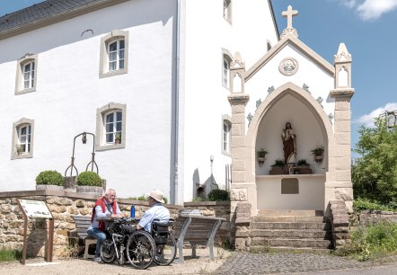 Zwei Männer, einer im Rollstuhl, sitzen auf einer Bank vor einem weißen Gebäude mit einer religiösen Statue in einer Nische. Sonniger Tag., © Naturpark Südeifel, Thomas Urbany Zwei Männer, einer im Rollstuhl, sitzen auf einer Bank vor einem weißen Gebäude mit einer religiösen Statue in einer Nische. Sonniger Tag., © Naturpark Südeifel, Thomas Urbany