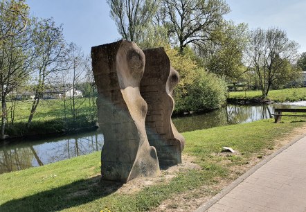 Stenen sculptuur aan de oever van de rivier, omgeven door groen landschap en bomen. Een heldere hemel en een pad zijn ook zichtbaar., © TI Bitburger Land, Steffi Wagner Stenen sculptuur aan de oever van de rivier, omgeven door groen landschap en bomen. Een heldere hemel en een pad zijn ook zichtbaar., © TI Bitburger Land, Steffi Wagner