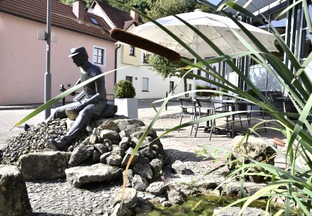 Bronzestatue eines Mannes auf Steinen, umgeben von Schilf und einem kleinen Teich. Im Hintergrund sind Häuser und ein Sonnenschirm zu sehen., © Tourist-Information Bitburger Land Bronzestatue eines Mannes auf Steinen, umgeben von Schilf und einem kleinen Teich. Im Hintergrund sind Häuser und ein Sonnenschirm zu sehen., © Tourist-Information Bitburger Land
