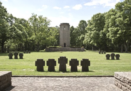 Ein Kriegsdenkmal mit Steinkreuzen auf einer Wiese, ein Turm im Hintergrund, umgeben von grünen Bäumen unter blauem Himmel., © TI Bitburger Land Ein Kriegsdenkmal mit Steinkreuzen auf einer Wiese, ein Turm im Hintergrund, umgeben von grünen Bäumen unter blauem Himmel., © TI Bitburger Land