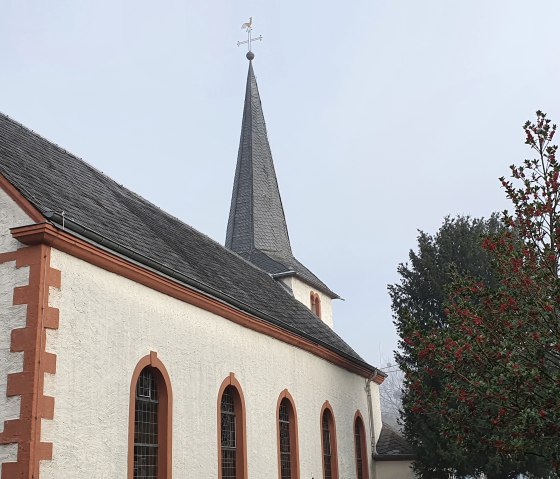 Church with red border and cross on the façade, next to it a tree with red berries., © TI Bitburger Land - Steffi Wagner