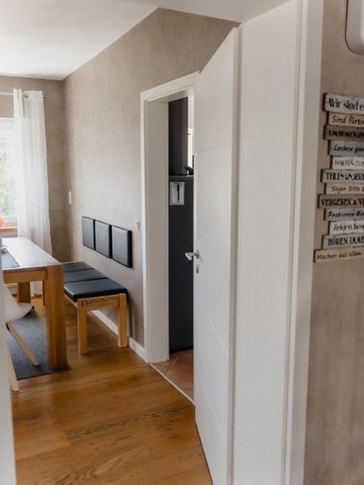 Ein heller Flur mit einem offenen Raum, der einen Tisch und Stühle zeigt. An der Wand hängen ein paar Bilder und es gibt eine offene Tür zu einem weiteren Raum.