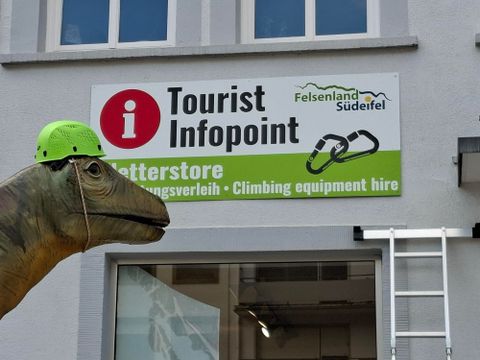 Een toeristeninformatiepunt met een bord dat klimuitrusting aanbiedt. Op de voorgrond staat een groot dinosaurussculptuur met een groene helm.