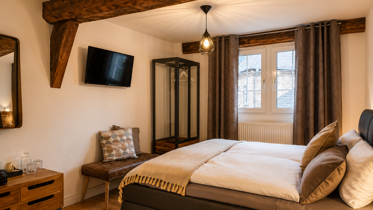 Een mooie slaapkamer met een groot bed en een gezellige stoel. De houten balken en de televisie geven de ruimte een uitnodigende sfeer.