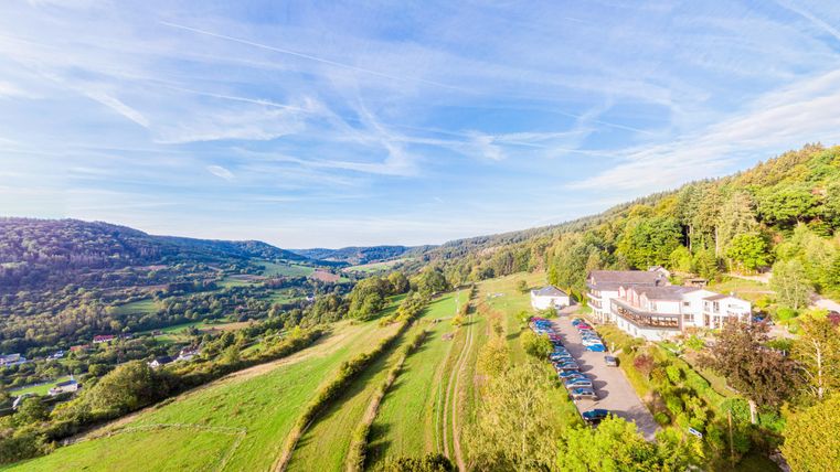 Het schilderachtige landschap met zachte heuvels en weelderig groen. Op de voorgrond staat het Boshotel Sonnenberg, omgeven door bomen en een parkeerplaats.