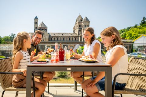 Familie mit zwei Kindern sitzt bei sonnigem Wetter auf der Terrasse einer Gaststätte und genießt ein gemeinsames Essen. Im Hintergrund die eindrucksvolle romanische Basilika Maria Laach mit ihren markanten Türmen. Auf dem Tisch stehen bunte Getränke und verschiedene Speisen. Die Stimmung ist fröhlich und entspannt.