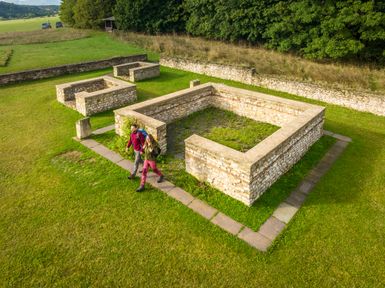 Luftaufnahme des Matronenheiligtums Görresburg bei Nettersheim. Zu sehen sind die freigelegten Grundmauern römischer Kultbauten aus Stein, eingebettet in eine grüne Wiese. Zwei Besucher Wandern vorbei an den zentralen Strukturen. Im Hintergrund verläuft eine Baumreihe entlang einer Mauer.