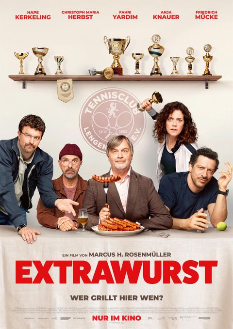 Ein Filmplakat für "Extrawurst" mit mehreren Personen an einem Tisch. Sie lächeln, während einer eine Wurst hält, und umgeben von Trophäen.