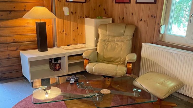A cozy living area with a comfortable chair and a glass table. The walls are made of wood and there is a warm glow from a lamp.