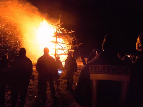 Eine Gruppe von Menschen beobachtet ein großes Feuer, das in der Nacht brennt. Im Hintergrund ist eine Holzstruktur zu sehen, die in Flammen steht.