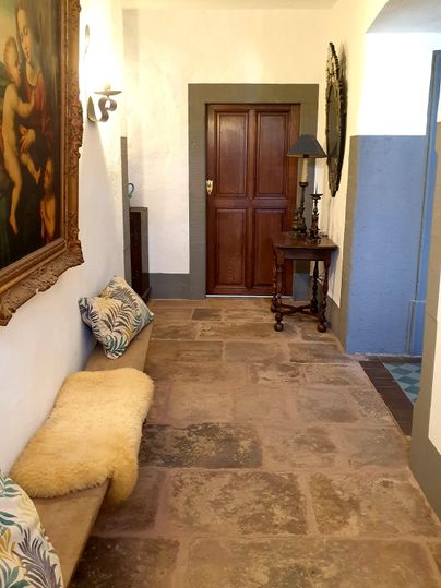 An elegant hallway with a painting on the wall and a wooden chair. The floor is laid with large stone tiles and the walls are painted in a light color.