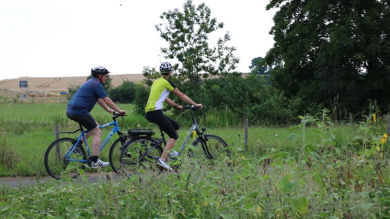 Two people ride bicycles on a path through a green landscape.