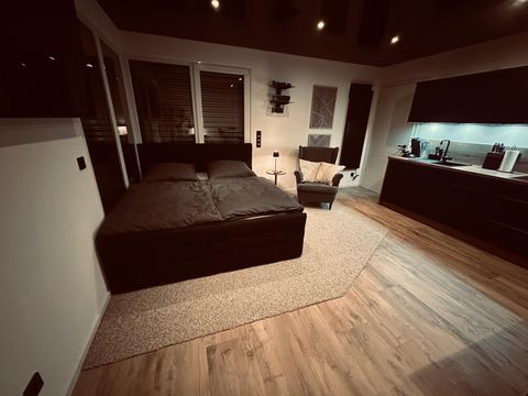 A modern bedroom with a large bed, a comfortable armchair and an elegant kitchenette. The atmosphere is cozy and stylish, with warm light and wooden flooring.