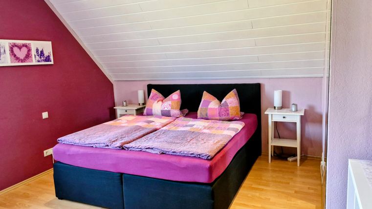 Een gezellige slaapkamer met een groot bed en kleurrijke kussens. De muren zijn in zachte kleuren geschilderd en er staan kleine nachtkastjes naast.