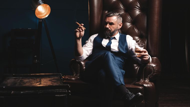 Ein stilvoller Mann sitzt in einem großen Lehnsessel und hält ein Glas Whisky in der Hand. Er trägt einen Anzug und raucht eine Zigarre, während er entspannt wirkt.