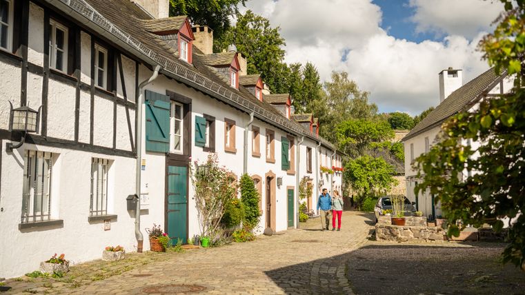 Maisons historiques à colombages dans une rue pavée de Kronenburg, entourées d'arbres et de plantes à fleurs.
