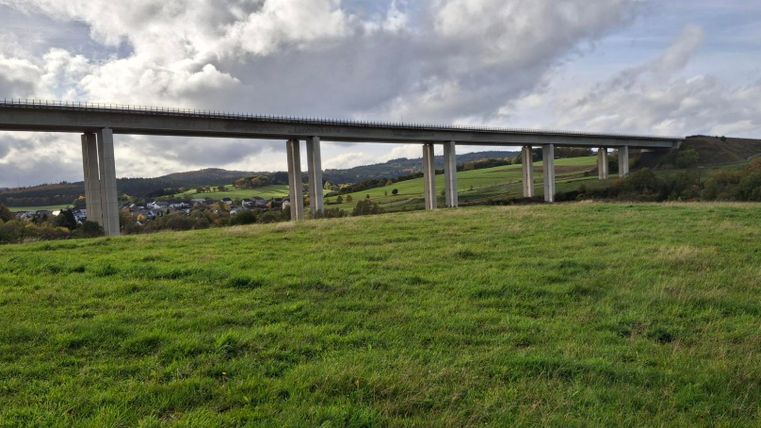Een grote betonnen brug die over een groene weide leidt. Op de achtergrond zijn zachte heuvels en een bewolkte lucht te zien.
