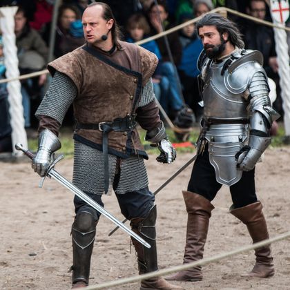 Zwei Ritter in historischem Kostüm stehen in einem Kampfgelände. Der eine trägt Leder- und Kettenrüstung, der andere eine glänzende Rüstung.