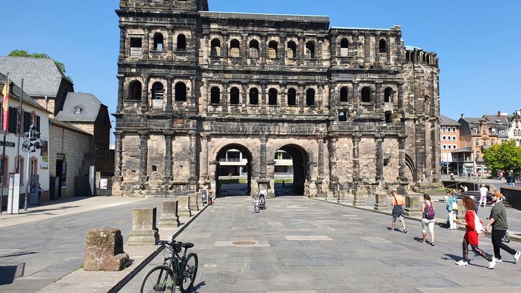 Porta Nigra in Trier, Duitsland, bij zonnig weer met voorbijgangers en een fiets op de voorgrond.