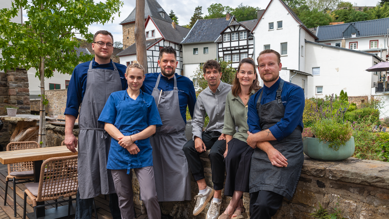 Six chefs se tiennent souriants devant un arrière-plan de bâtiments historiques. C'est une journée ensoleillée et l'ambiance est accueillante.