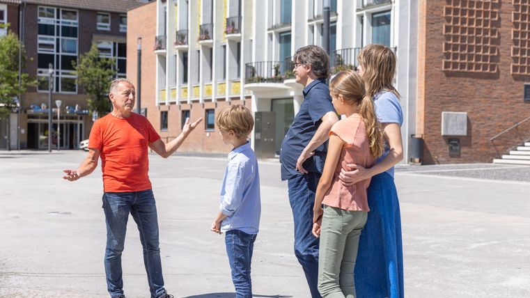 Eine Gruppe von Menschen steht im Freien und spricht miteinander. Im Hintergrund sind moderne Gebäude zu sehen.