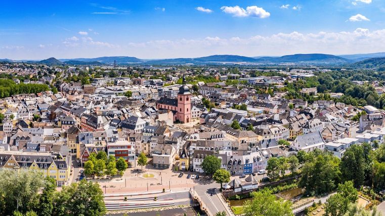 Une vue pittoresque de la ville avec de nombreuses maisons et une tour marquante. L'environnement est vert et vallonné, sous un ciel bleu clair.
