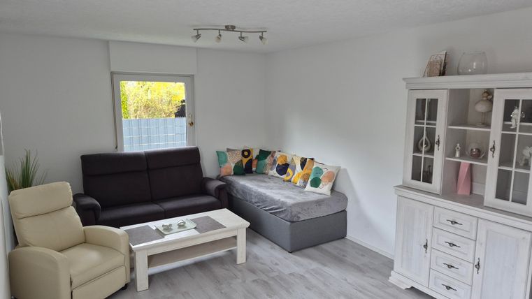 Een lichte woonkamer met een comfortabele bank en een stoel. Een decoratieve kast staat tegen de muur, en het licht valt door het raam.