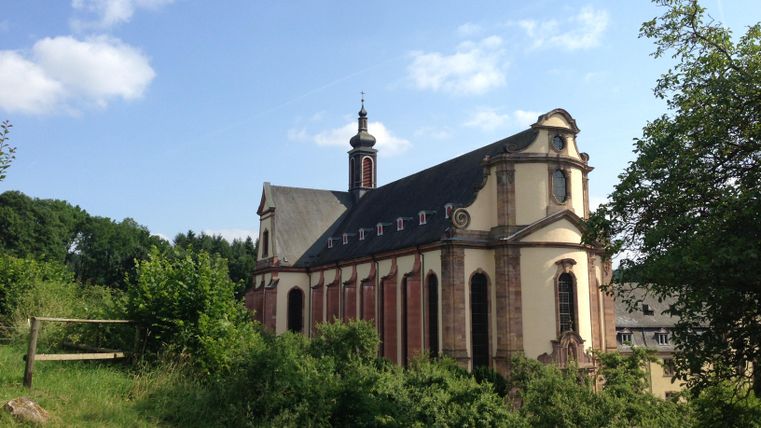 Een historische kerk omringd door groene weilanden en bomen. De lucht is blauw met enkele wolken.