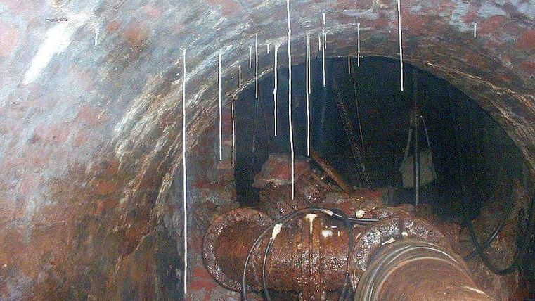 Een blik in een donkere tunnel met rode bakstenen muren. Aan de wanden hangen waterdruppels en er zijn buizen te zien.