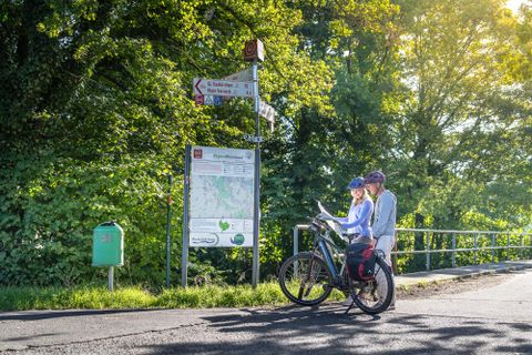 Twee fietsers staan bij een wegwijzer en bekijken een kaart. Op de achtergrond zijn groene bomen en zonnig weer te zien.