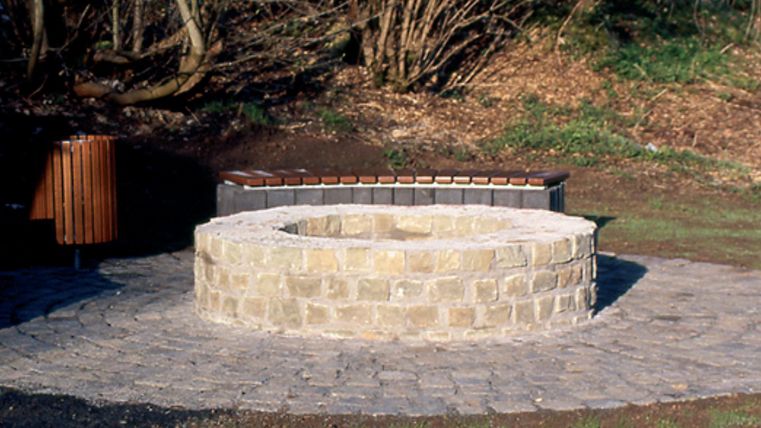 Runder, steinerner Brunnen mit Bank und Mülleimer im Hintergrund, umgeben von Bäumen.