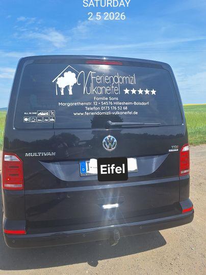Ein schwarzer Volkswagen Multivan mit einem Werbeaufdruck für eine Ferienunterkunft. Im Hintergrund ist ein klarer Himmel zu sehen.