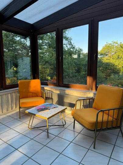 Ein gemütlicher Wintergarten mit zwei gelben Stühlen und einem kleinen Tisch. Im Hintergrund ist die grüne Natur sichtbar.