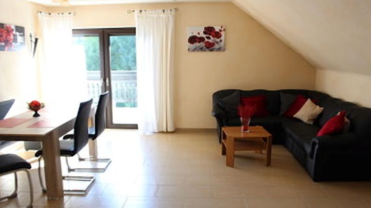 A modern living room with a cozy sofa, a large TV, and a dining area. Through the balcony doors to the east balcony, the light of the rising sun floods the room and creates a bright atmosphere.