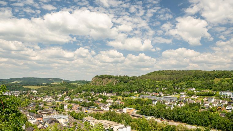 Blick von oben auf die Stadt Gerolstein mit zahlreichen Gebäuden. DIe Stadt ist von hohen Felsen und Bäumen umgeben. Am blauen Himmel hängen viele kleinen Wolken.