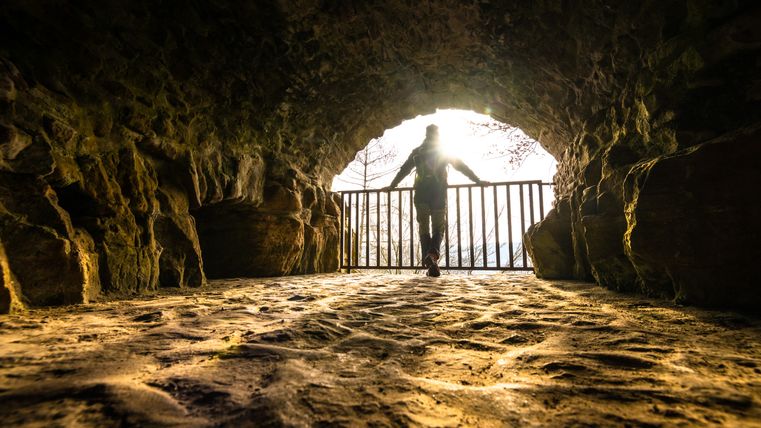 Persoon staat in een stenen tunnel, verlicht door gouden licht, met uitzicht op een getraliede poort.