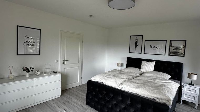 Ein modernes Schlafzimmer mit einem großen, gepolsterten Bett und hellen Wänden. Der Raum ist schlicht eingerichtet und hat einen eleganten Teppich sowie Dekorationen an den Wänden.