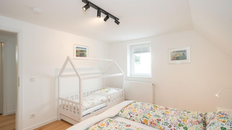 Ein helles Schlafzimmer mit einem Doppelbett und einem Kinderbett in Form eines Hauses. Die Wände sind hell gehalten und mit Bildern dekoriert.