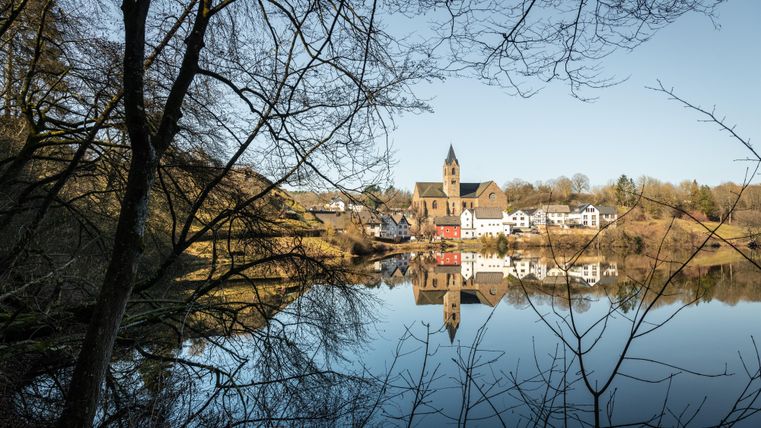 Een rustig landschap met een helder meer waarin de kerk en de omliggende gebouwen weerspiegeld worden. Op de achtergrond zijn bomen en een heldere hemel te zien.