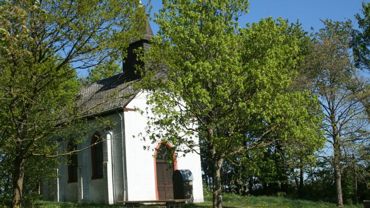 Une petite chapelle blanche entourée d'arbres et d'une prairie sous un ciel bleu.