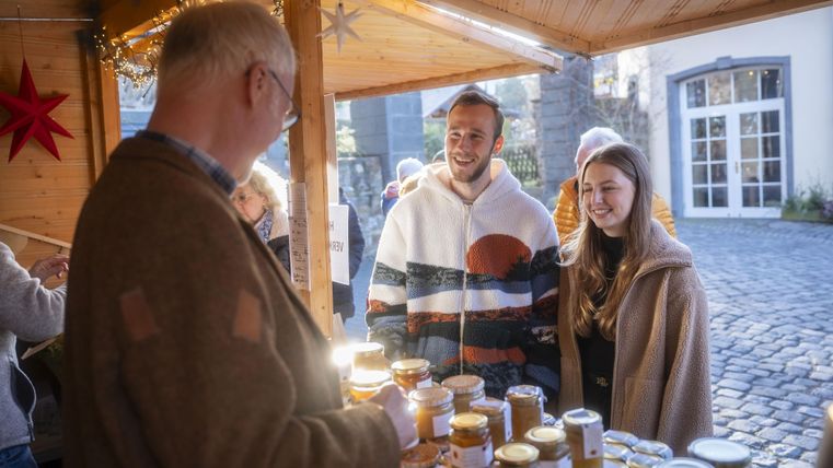 Ein lebhafter Marktstand mit einem Verkäufer, der mit zwei jungen Kunden spricht. Auf dem Tisch sind viele Gläser mit verschiedenen Produkten zu sehen.