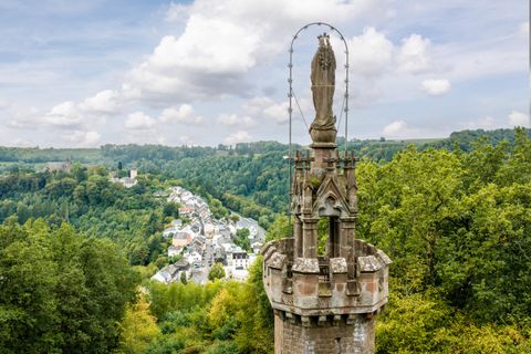 Eine Statue auf einem hohen Turm blickt über eine grüne Landschaft und ein kleines Dorf im Tal. Der Himmel ist teilweise bewölkt und die Natur ist üppig.