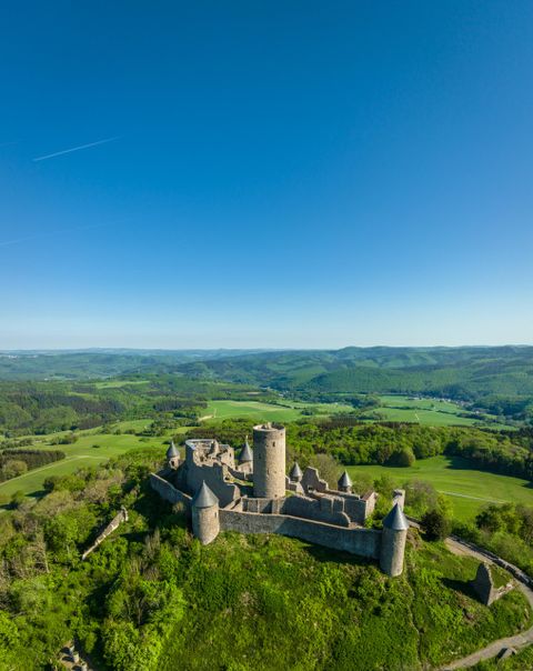 Eine beeindruckende Burgruine auf einem hügeligen Gelände. Rundherum erstrecken sich grüne Wiesen und ein klarer blauer Himmel.