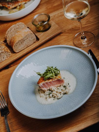 Een bord met gebraden vis op romige risotto, gegarneerd met verse rucola. Daarbovenop een glas witte wijn en vers brood op de tafel.