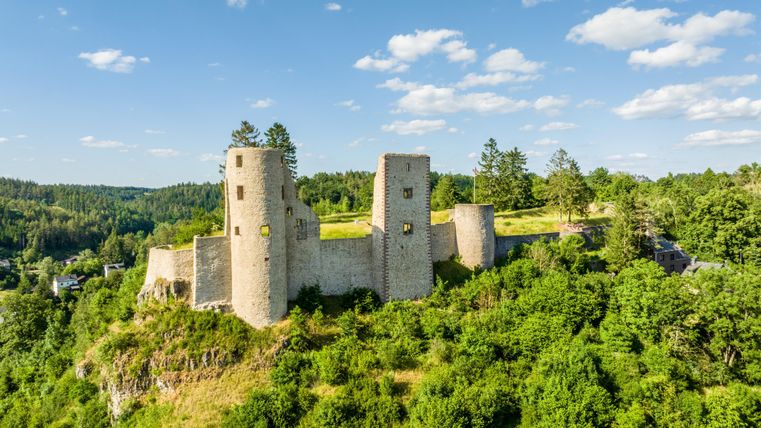 An old castle ruin on a hill, surrounded by green forests. The sky is clear with a few white clouds.