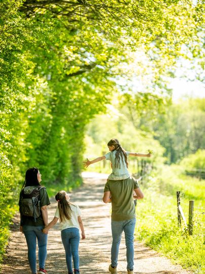 A family walks along a sunny path, surrounded by green trees. A child sits on a parent's shoulders while the others hold hands.