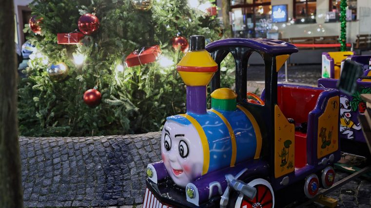 Een kleurrijke miniatuurtrein staat voor een feestelijk versierde kerstboom. Op de achtergrond zijn lichten en kerstdecoraties te zien.