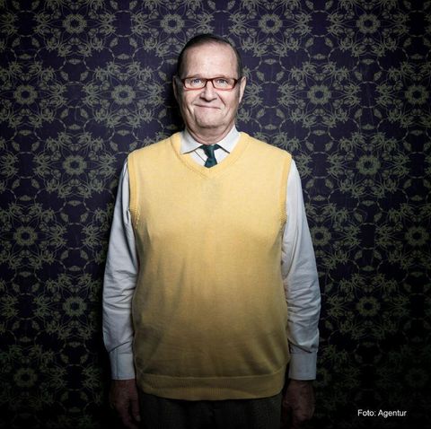 Een oudere man met een bril draagt een gele trui over een wit overhemd en een groene stropdas. De achtergrond is ingericht met een bloemmotief.